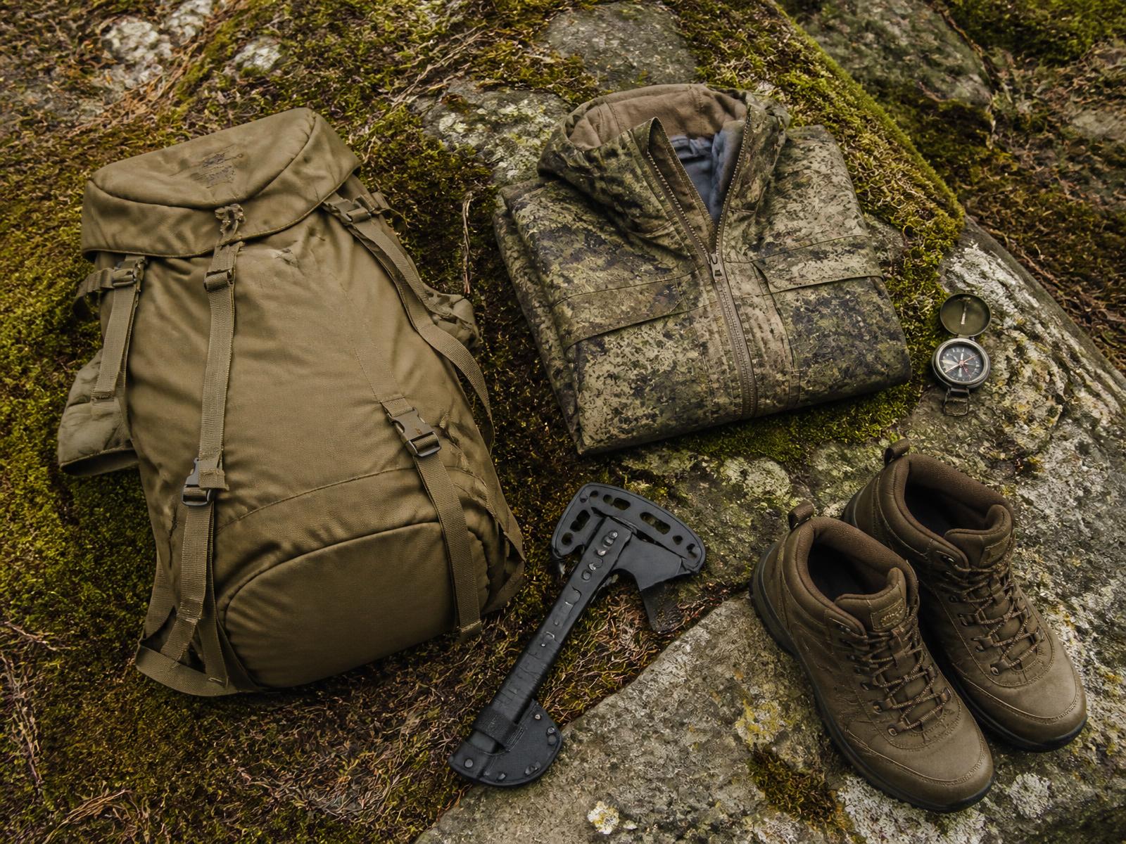 Outdoor equipment including a backpack, camouflage jacket, compass and boots arranged on mossy rock